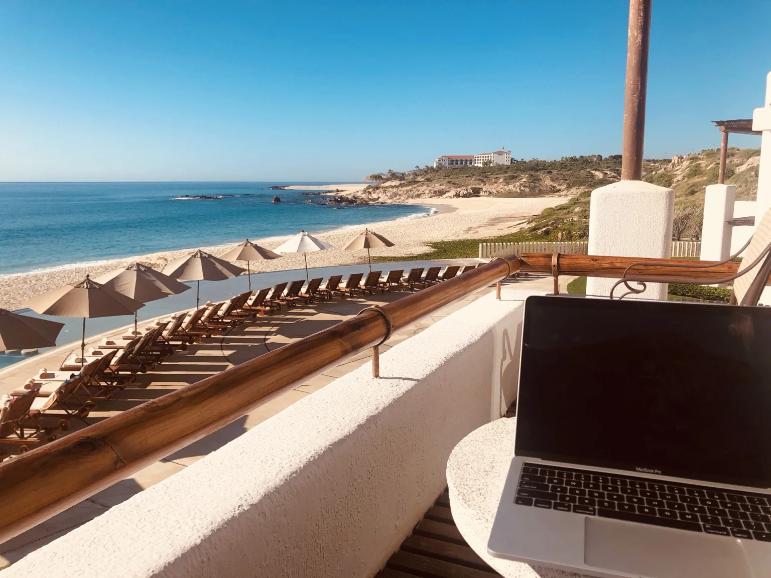 An open laptop on a balcony table overlooking the beach and pool.